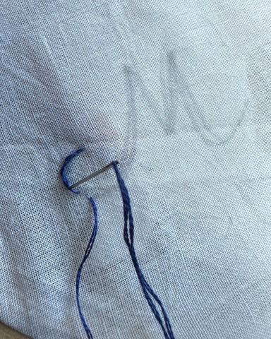Letter M on white fabric, first stitches of stem stitch embroidery with blue thread.