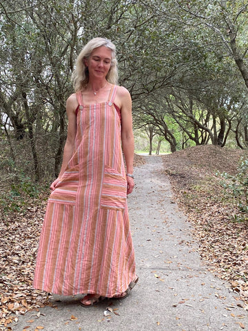 Woman standing in a forest wearing a striped peach colored long pinafore dress.