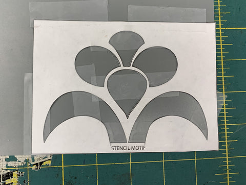 Floral design cut out of white paper and taped to a grey sheet of plastic stencil paper. On a green cutting mat.