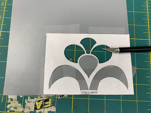 Floral design cut out of white paper and taped to a grey sheet of plastic stencil paper with pieces cut out of the stencil paper. On a green cutting mat, with a exacto knife on the right.