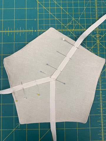 Pinned three beige ribbons on the top layer of biscuit cozy pattern in the shape of a pentagon cut out of a beige linen on a green cutting mat.