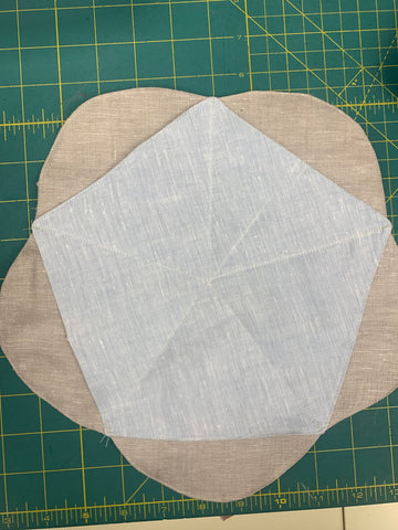 Stitched wrong the side of MIDDLE LAYER pentagon in light blue linen fabric to right side of BOTTOM LAYER scalloped piece in a dark beige linen fabric from center to corner of MIDDLE LAYER, matching Stitching Lines on a green cutting mat.