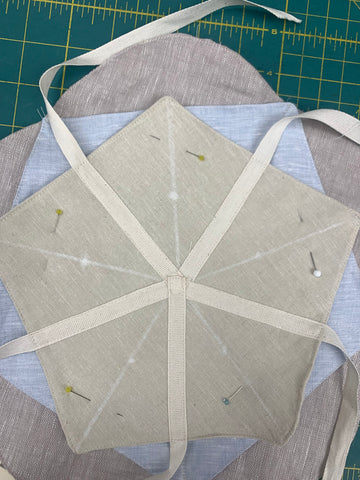 Pinned wrong side of the TOP LAYER pentagon in light beige with the ribbons already sewn on to right side of the MIDDLE LAYER pentagon in light blue only. DO NOT catch in BOTTOM LAYER and stitch along the stitching lines from small dots to corners of TOP LAYER. On a green cutting mat.
