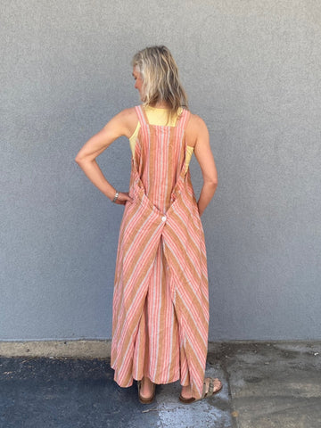 woman standing in front of a grey wall wearing a peach colored striped long pinafore dress - her back is to the camera.