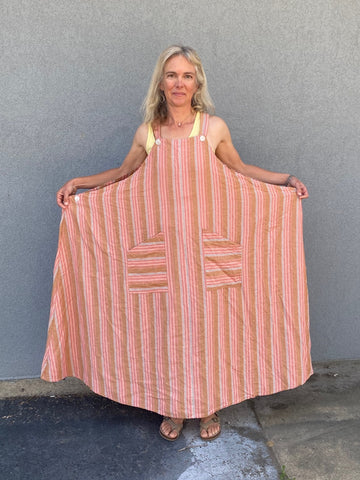 woman standing in front of a grey wall wearing a peach colored striped long pinafore dress - holding the sides out to show the expanded version