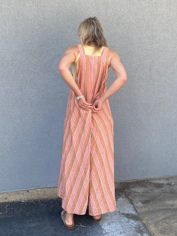 woman standing in front of a grey wall wearing a peach colored striped long pinafore dress - her back is to the camera and she is showing how to button the dress in the back.