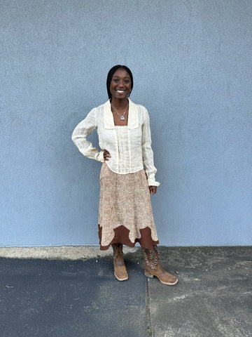 Woman standing outside by a grey wall wearing a brown western skirt and cream long sleeve blouse.