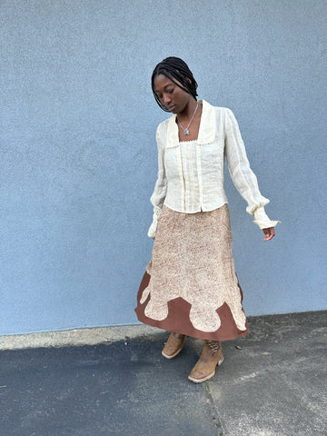 African American Woman wearing a silk floral beige and brown rodeo cowgirl skirt with brown arrowhead hem flounce, wearing brown cowgirl boots and 210 armistice blouse, looking down. She is standing in front of a light grey blue wall outside.