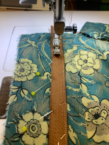 Sewing machine foot sewing right side of pinned center of the zipper teeth onto the right side of the facing D. On teal and taupe floral fabric.