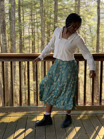 African American Woman twirling towards her right, wearing 210 armistice blouse in a light beige and  209 walking skirt in floral teal/taupe viscose fabric. On a porch outside.