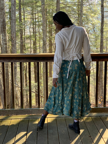 The back of an African American Woman wearing 210 armistice blouse in a light beige and  209 walking skirt in floral teal/taupe viscose fabric. Standing on a porch outside left leg out looking down towards her left.