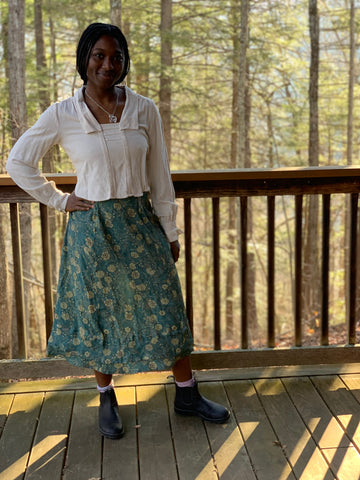A smiling African American Woman standing with her right hand on her hip, wearing 210 armistice blouse in a light beige and  209 walking skirt in floral teal/taupe viscose fabric. On a porch outside.