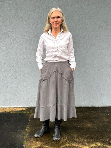 Woman standing outside wearing a grey skirt and white button down shirt with black boots.  Her hands are in her pockets.