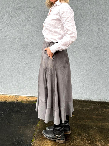 Woman standing outside wearing a grey skirt and white button down shirt with black boots - side view.  Her hands are in her pockets.