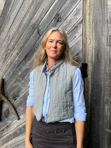 woman standing in front of a wooden door wearing a short grey vest and blue shirt