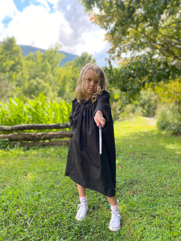 A young girl is wearing a black dress and is pointing a witch's wand at the viewer