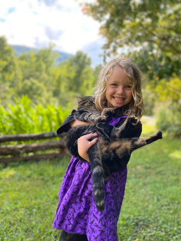 A young girl is wearing a purple apron and black dress, and is holding a cat