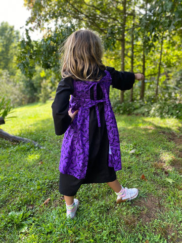 A young girl is wearing a black dress with a purple apron and is holding a witch's wand. This photo shows the back of the apron, which ties together.