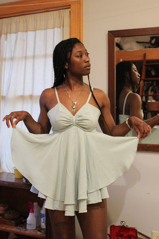 African American woman looking to her left, wearing a aqua light blue Juliette's Dream garment, in a room with her reflection in a mirror on a wall on the right, holding layered skirt out with both hands.