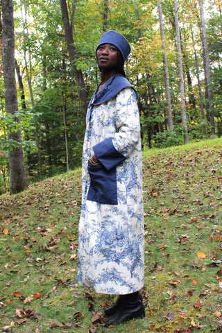 African American Woman wearing 262 spectators coat with hands in pockets turned towards her right. The coat is made with a navy voile for the collar, cuffs and pockets with the main fabric out of a french toile cotton canvas navy and off white. She is also wearing a Cloche hat with a navy blue brim out of the navy cotton voile. She is standing outside in the woods.