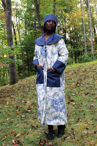 African American Woman wearing 262 spectators coat with hands in pockets. The coat is made with a navy voile for the collar, cuffs and pockets with the main fabric out of a french toile cotton canvas navy and off white. She is also wearing a Cloche hat with a navy blue brim out of the navy cotton voile. She is standing outside in the woods.