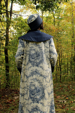 African American Woman wearing 262 spectators coat showing the back of the coat. The coat is made with a navy voile for the collar, cuffs and pockets with the main fabric out of a french toile cotton canvas navy and off white. She is also wearing a Cloche hat with a navy blue brim out of the navy cotton voile. She is standing outside in the woods.