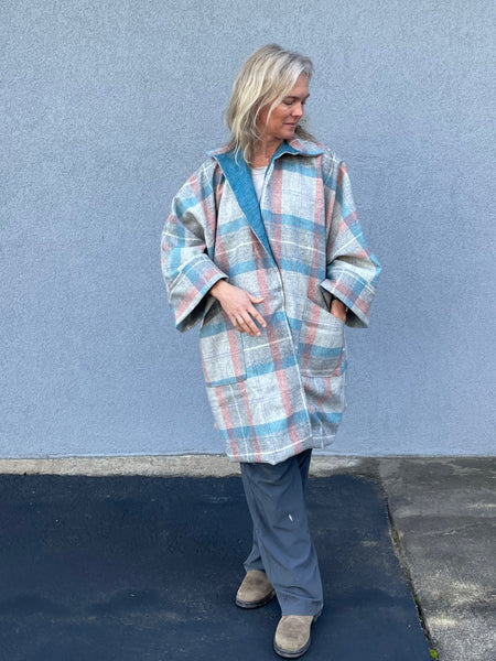 Woman standing outside by a grey wall in a plaid overcoat