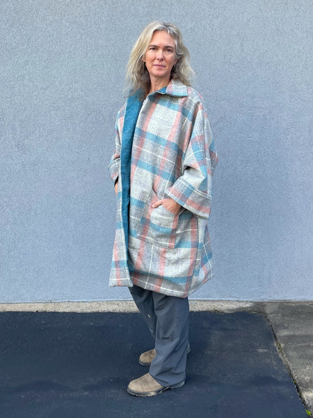 Woman standing outside by a grey wall in a plaid overcoat