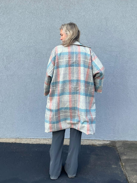 Woman standing outside by a grey wall in a plaid overcoat, back toward camera.