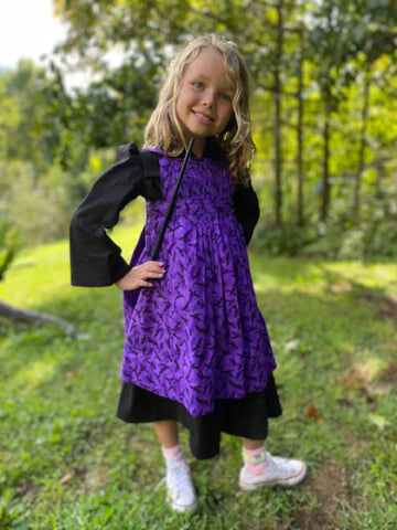 Young girl in a black dress with a purple pinafore over it that has black bats.  She is standing outside holding a wooden wand.