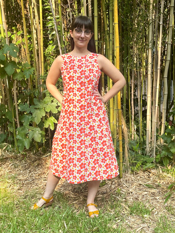 Woman standing in front of bamboo forest in an orange floral muumuu, wearing yellow shoes.