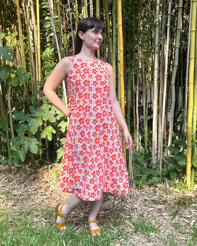 Woman standing in front of a bamboo forest with one hand on hip, wearing an orange floral muumuu and yellow sandals.