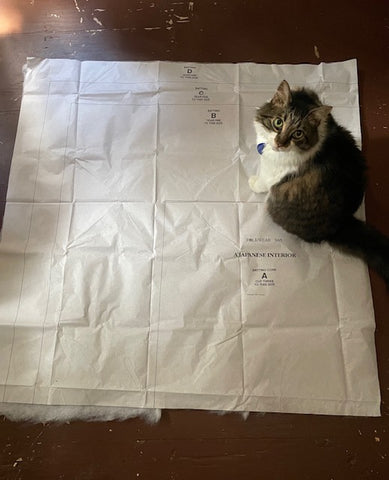 A fluffy cat is sitting on a sewing pattern tissue page on the floor