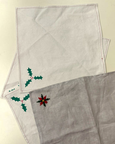 Three napkins, two painted with holly leaves and one embroidered with a poinsettia on cream and grey fabrics.