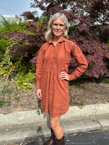 Woman standing in a burnt orange linen dress by a curve, with her left hand on her hip.