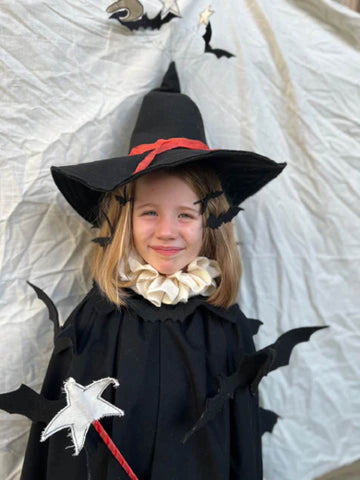 Young girl in a black witch's hat and a black coat with a white neck ruff, and black felt bats around her shoulders.  She is holding a star wand.