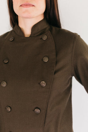 Close up of Woman standing wearing a brown Belgian Military Chef's Coats.  White backdrop.