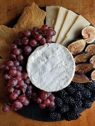 overhead view of white bloomy rind cheese on black slate cheese board with grapes, figs, crackers, blackberries, hard cheese