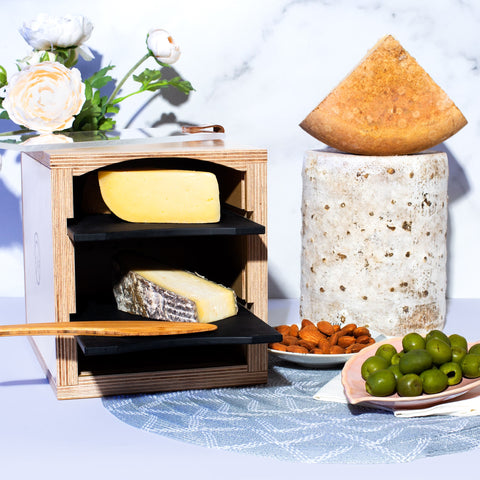 wooden cheese storage box holding wedges of cheese with wheels of cheese, olives, and charcuterie on table with flowers and marble background