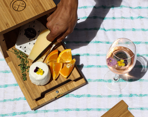 overhead view of hand with brown skin tone cutting into cheese on cheese board in cheese storage box against blue and white tablecloth with a glass of white wine