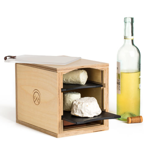 small cheese storage box with black slate sheles and bloomy rind cheese inside next to bottle of white wine against white background