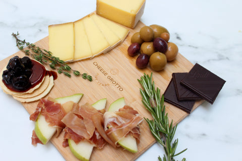 square bamboo cheese board with cheese, olives, charcuterie, chocolate, crackers, apples, and herbs