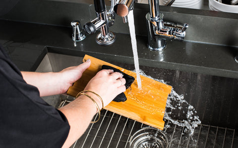 light skinned hands washing small bamboo cheese board at sink with sponge