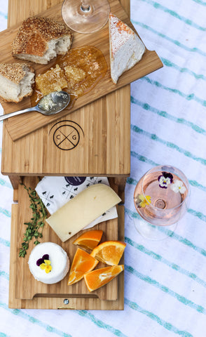 overhead view of bamboo cheese server sticking out of bamboo cheese aging box with cheese, fruit, and rose wine