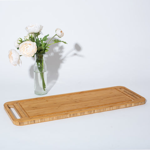 long, rectangular bamboo cheese server with carved handles on white background with glass vase of light pink flowers