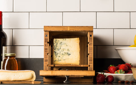 wooden cheese storage box with piece of blue cheese in it on countertop with wine bottle and strawberries