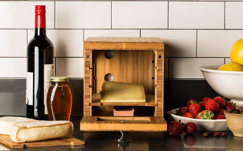 wooden cheese box with wedge of cheese surrounded by wine bottle, brie slice and strawberries on counter with white tile background