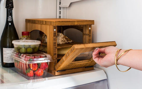 white person's hand with gold bracelets opening wooden cheese storage box in fridge with box of strawberries and bottle of wine 