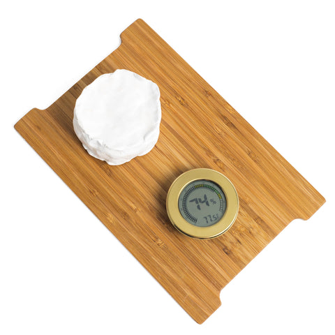 overhead view of small white bloomy rind cheese with gold digital hygrometer on bamboo cheese board against white background