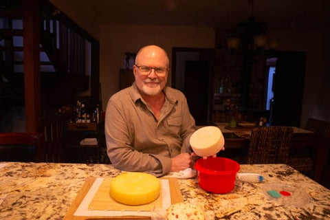 Fred Schenkelberg, home cheesemaker, in his kitchen with white, orange, and red wheels of cheese he made at home.